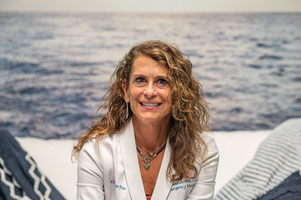 Dr. Paula Jones from Rejuvenate You in a white medical coat against an ocean backdrop (wide frame).