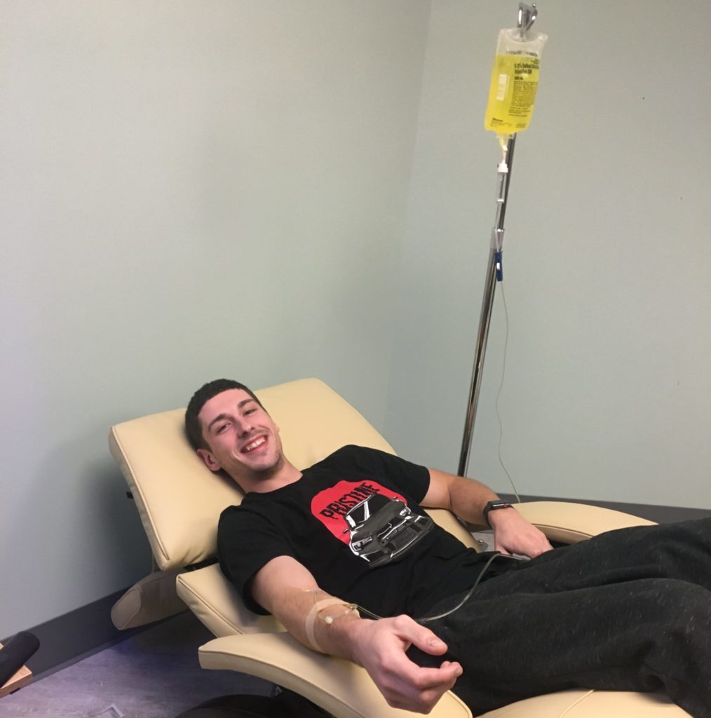 Young man smiling as he an IV bag drips nutrients into his right arm.