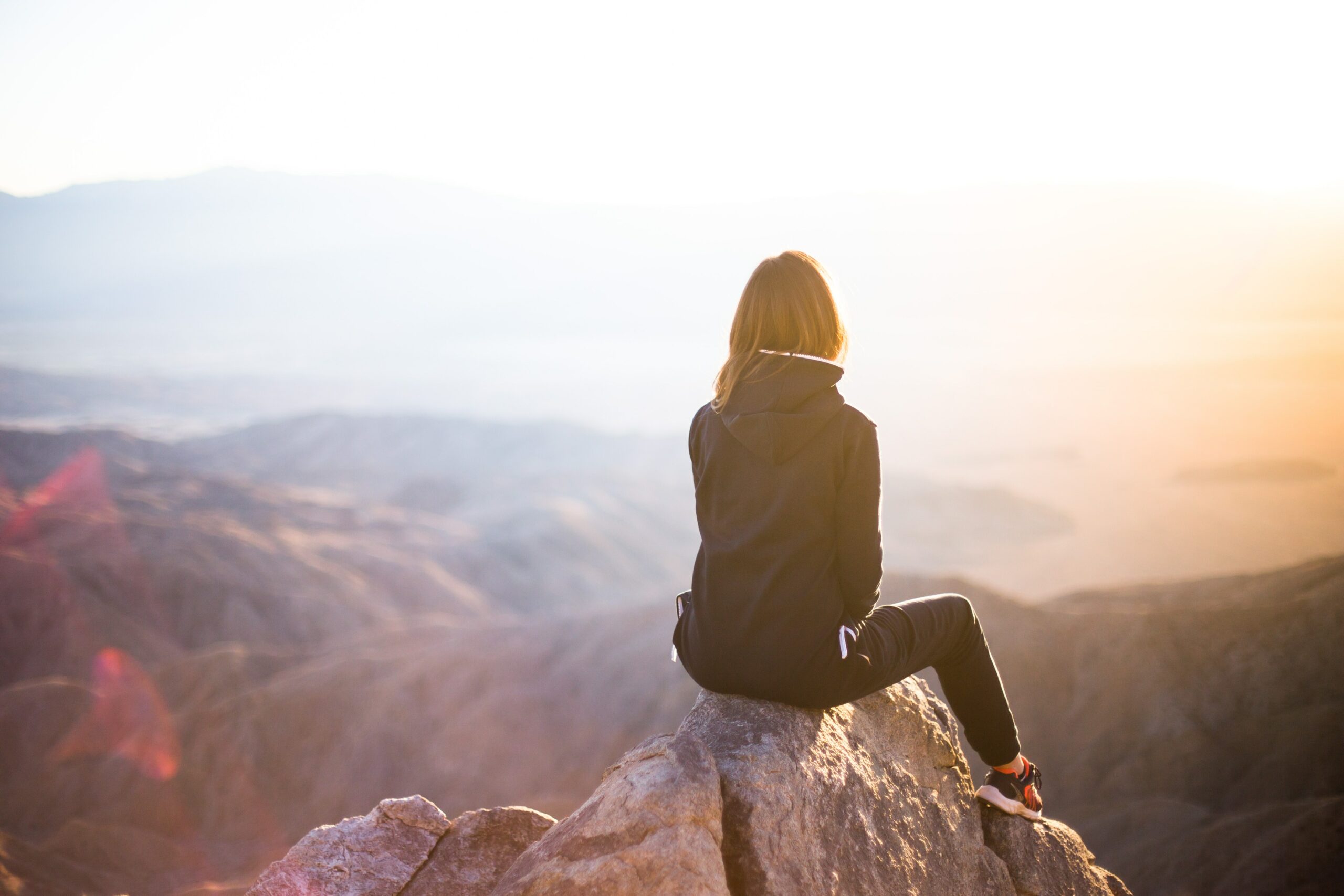 A woman sits on a rock facing out over a valley at sunset.