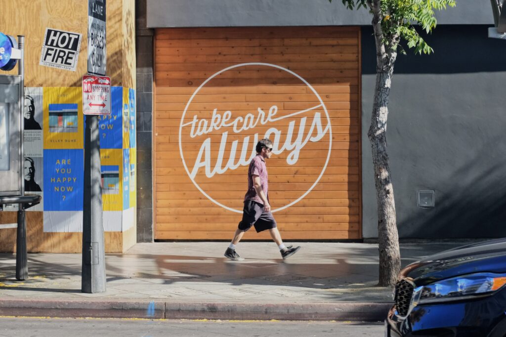 A fit man in sunglasses walking on a sidewalk in front of a building with the words "take care Always" painted on it in cursive.