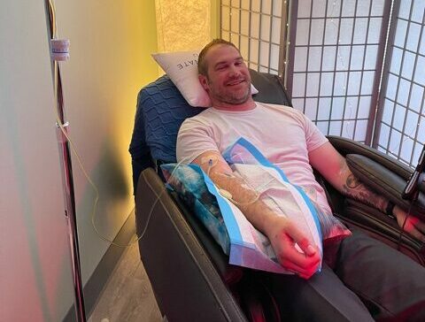 A tattooed and smiling man in a white t-shirt reclining with an crohn's disease infusion in his arm.