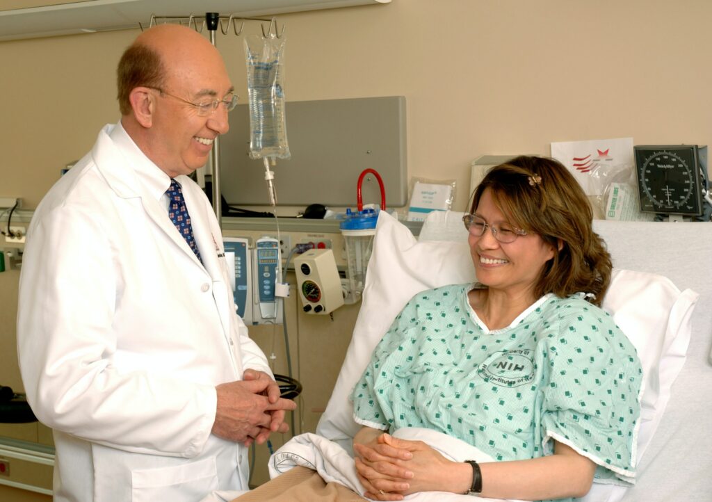 A smiling doctor gives an IV to a beaming woman in a green hospital gown.