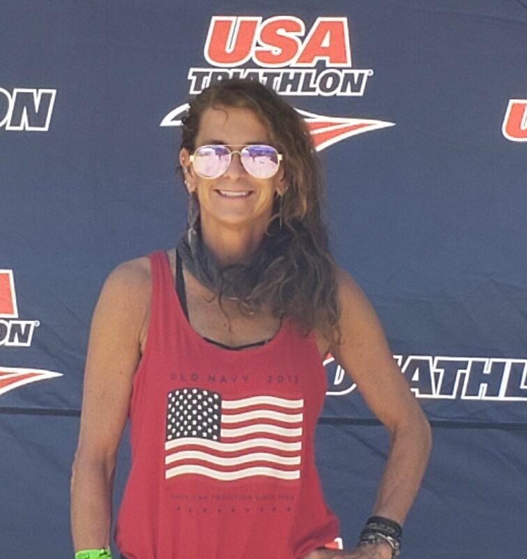 Paula Jones in sunglasses and a red American flag tank top in front of a USA Triathlon banner.