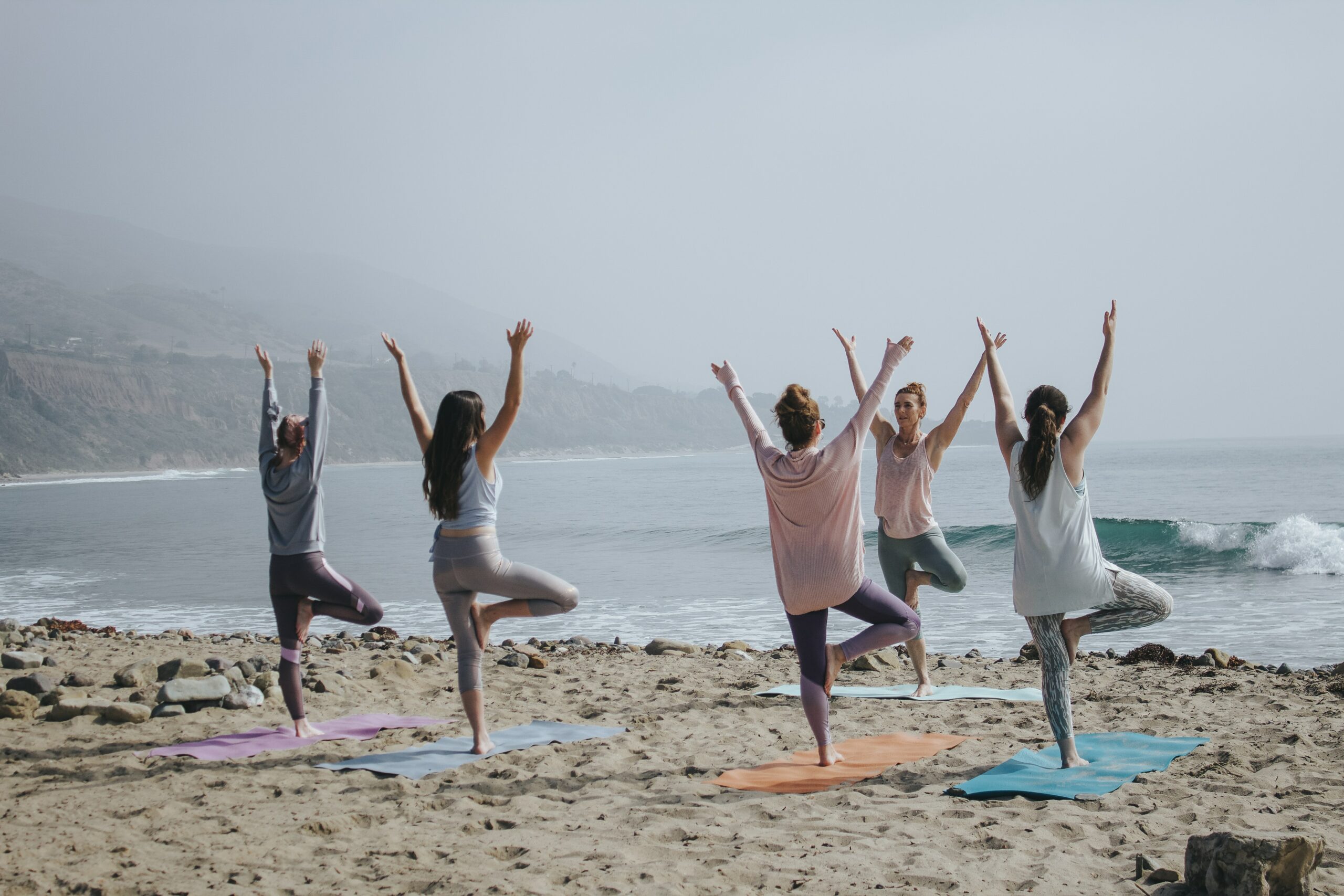 Five women standing on one leg each in yoga poses on a beach.