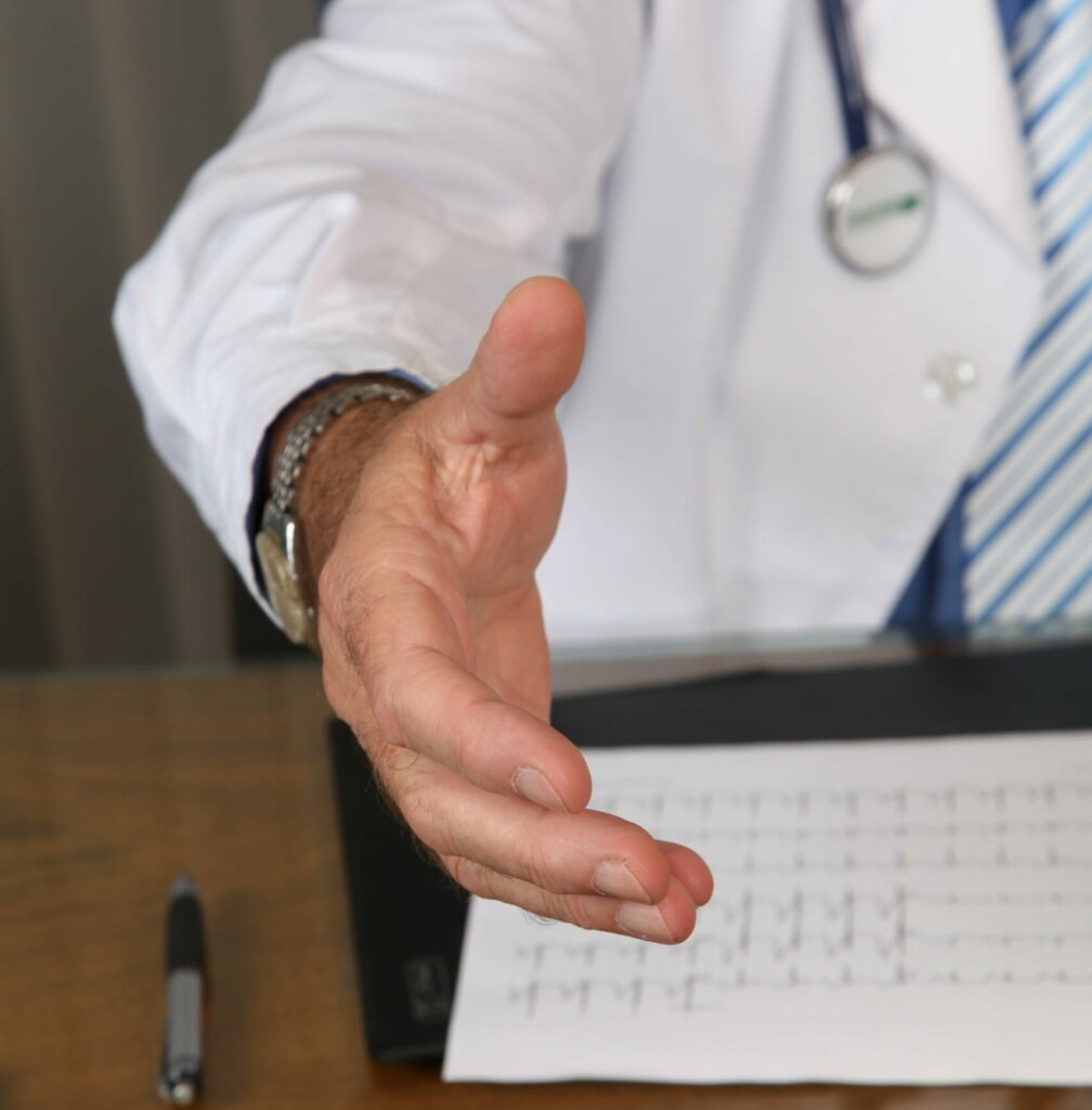 A male doctor's hand extended from where he sits behind a wooden desk.