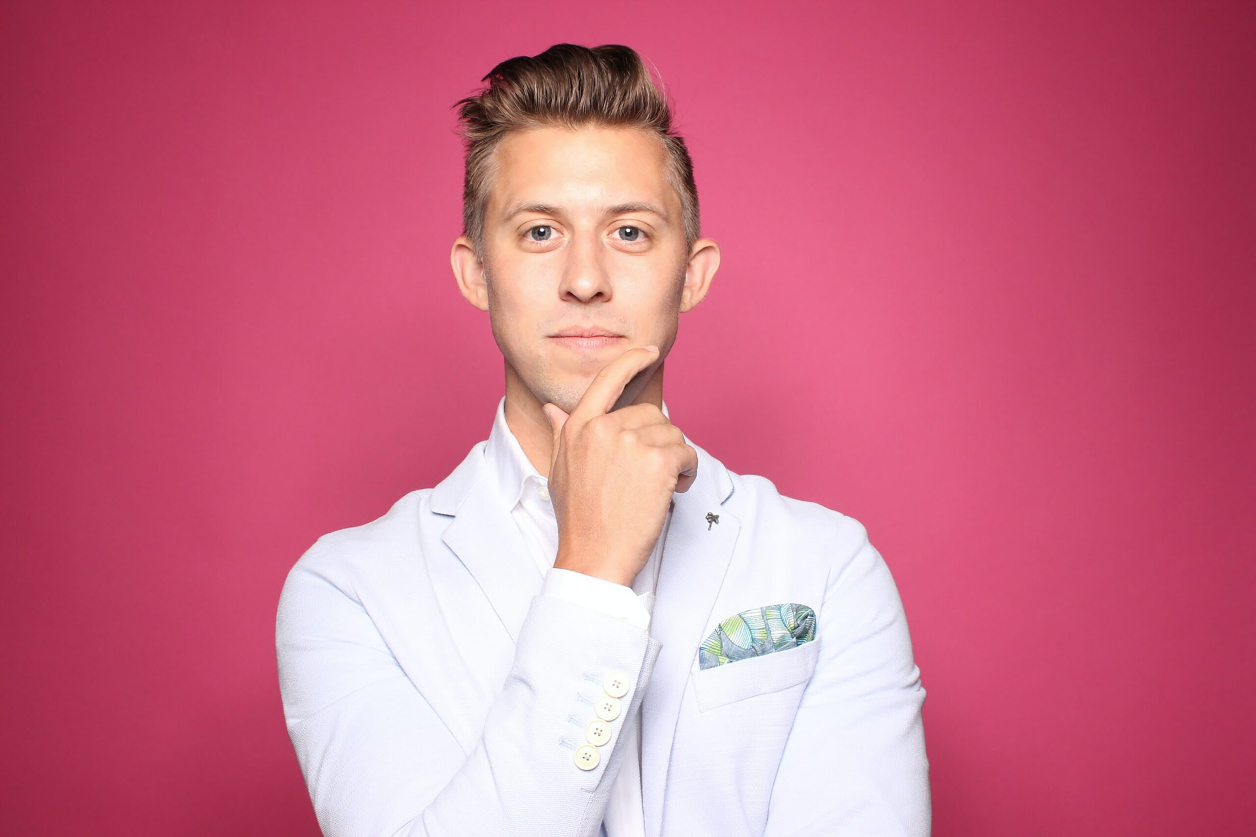 A man in a white suit stroking his chin in front of a rosy pink background.