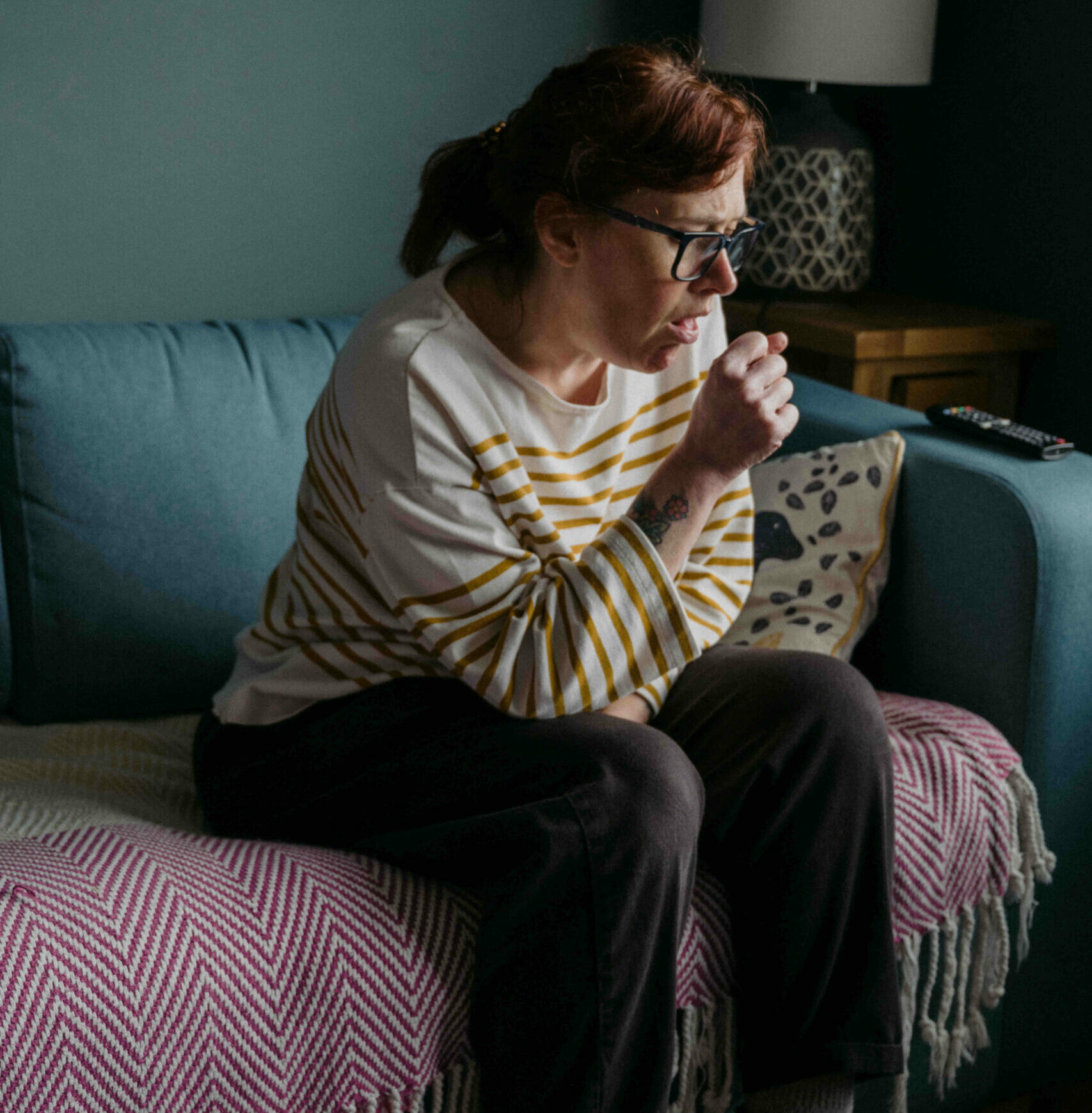 A woman sitting on a couch and coughing into her hand.