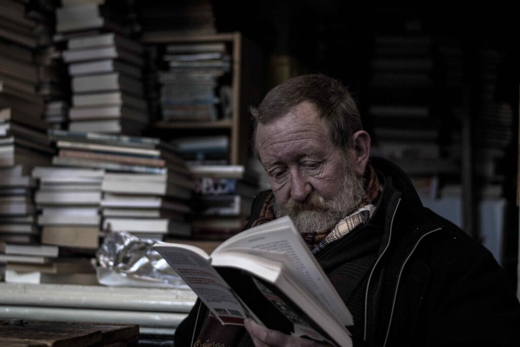 A bearded man reading beside many stacks of books.