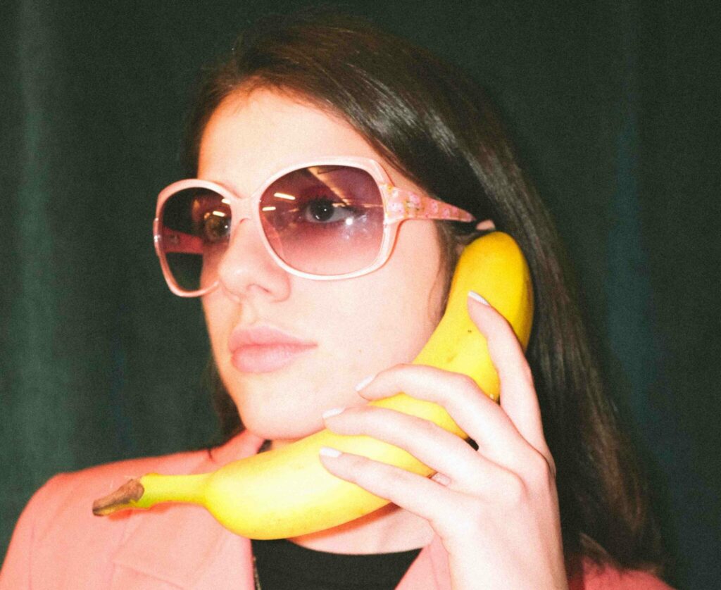 A woman in sunglasses holding a banana to her ear like it were a telephone.