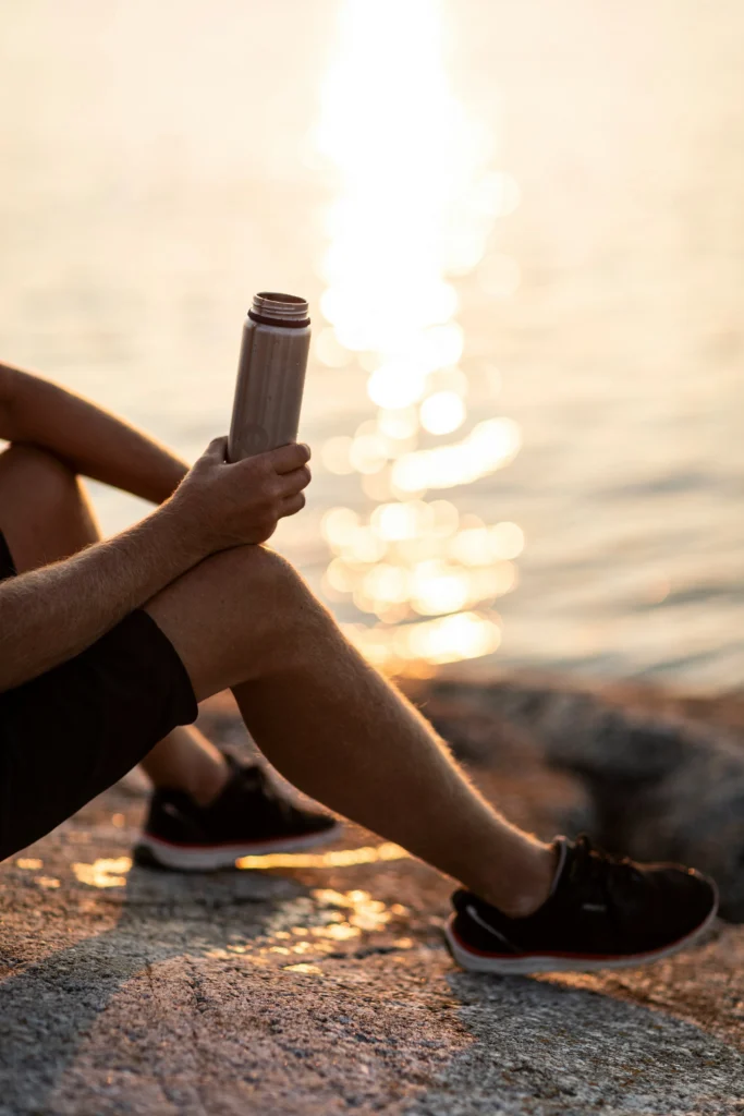 bluewater sweden vTEqYBgqcmM unsplash Person sitting by a body of water drinking from a water bottle.